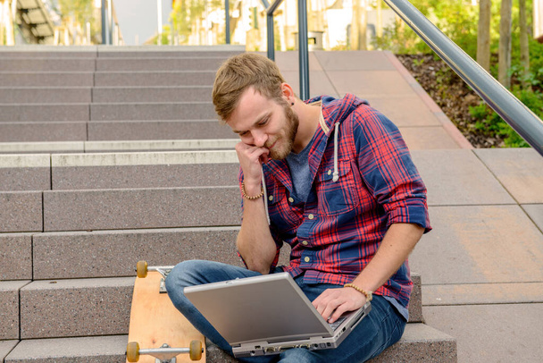 νεαρός άνδρας με καρό πουκάμισο κάθεται στις σκάλες και χρησιμοποιεί το laptop - Φωτογραφία, εικόνα