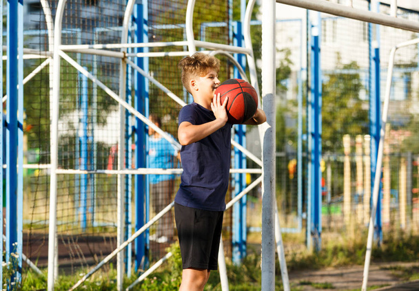 Bonito menino sorridente em azul t-shirt joga basquete no parque infantil da cidade. Adolescente ativo desfrutando de jogo ao ar livre com bola laranja. Hobby, estilo de vida ativo, esporte para crianças
. - Foto, Imagem