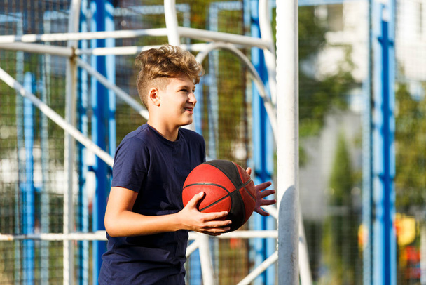 Cute smiling boy in blue t shirt plays basketball on city playground. Active teen enjoying outdoor game with orange ball. Hobby, active lifestyle, sport for kids. - Fotografie, Obrázek