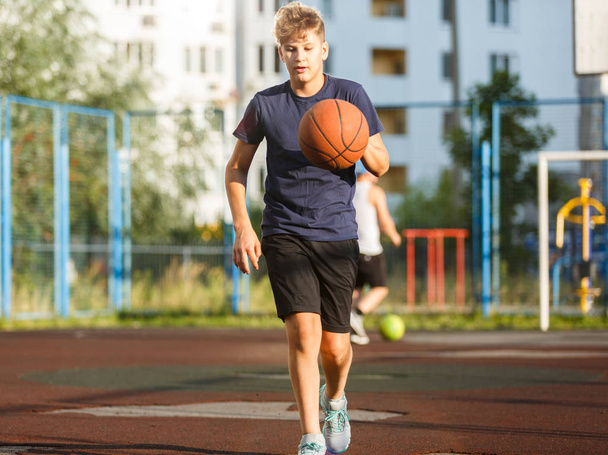 Cute smiling boy in blue t shirt plays basketball on city playground. Active teen enjoying outdoor game with orange ball. Hobby, active lifestyle, sport for kids. - 写真・画像