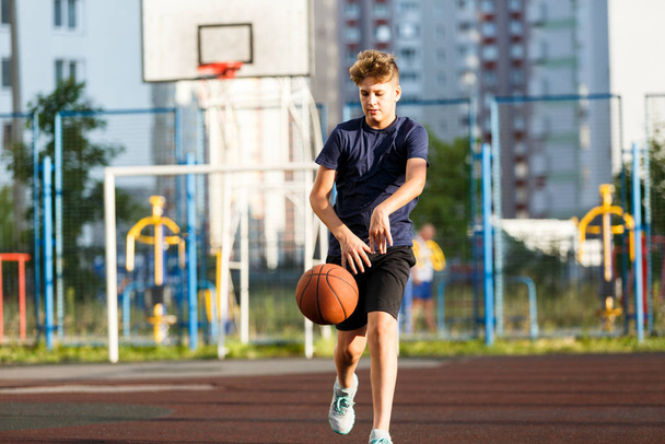 Cute smiling boy in blue t shirt plays basketball on city playground. Active teen enjoying outdoor game with orange ball. Hobby, active lifestyle, sport for kids. - Foto, imagen