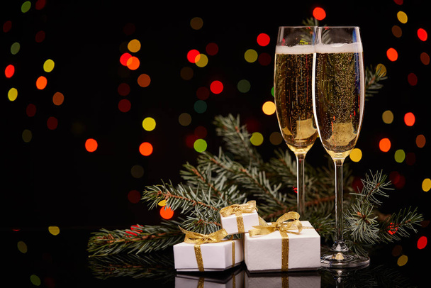 Two glasses with champange, fir tree branch, gift boxes on a black background with multi-colored lightes of garland.  New year and Christmas. - Foto, imagen