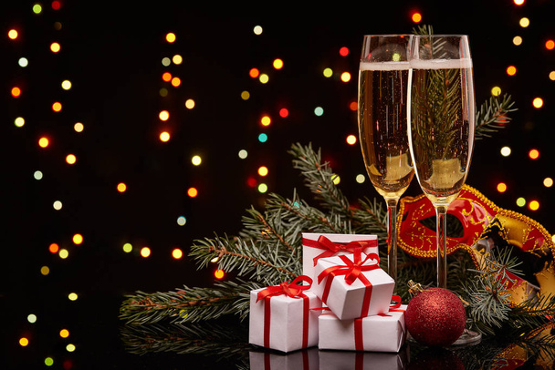 Two glasses with champange, fir tree branch with decoration, gift boxes on a black background with multi-colored lightes of garland.  New year and Christmas. - Photo, image
