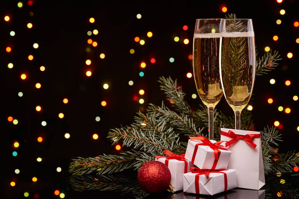 Two glasses with champange, fir tree branch with decoration, gift boxes on a black background with multi-colored lightes of garland.  New year and Christmas. - Foto, Imagem