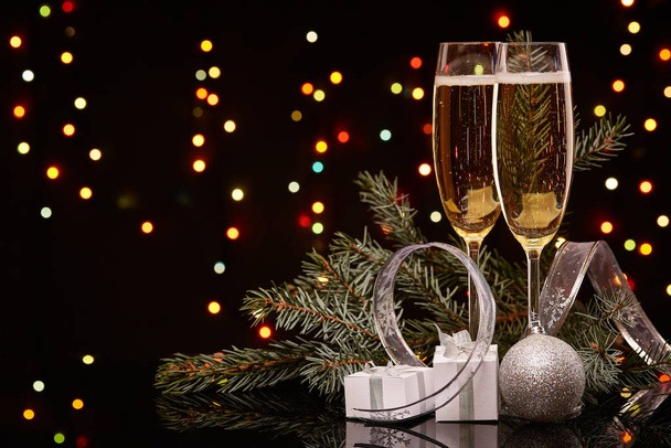 Два бокала с шампанским, елка с отделкой, подарочные коробки на черном фоне с разноцветными гирляндами. Новый год и Рождество. - Фото, изображение