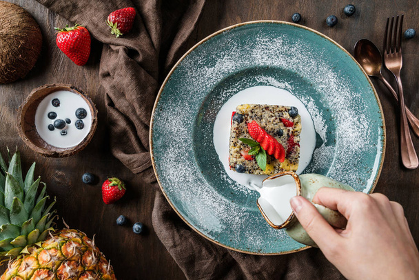 Hand pouring a coconut milk into quinoa porridge with fruits, strawberries, blueberry, pineapple over brown wooden background with linen napkin. Healthy vegan food, clean eating, dieting, top view - Photo, Image