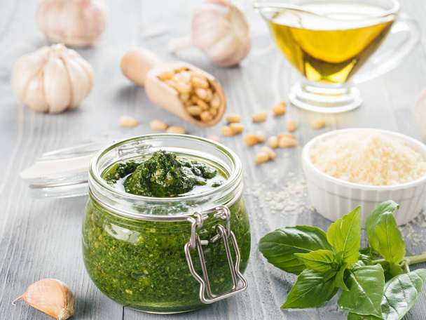 Homemade pesto sauce and ingredients on gray wooden background. Close up wiev of basil pesto in glass jar with ingredients. - Photo, image