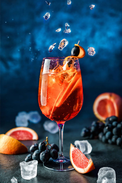 Cóctel de pomelo fresco con naranja, uvas y hielo en vino sobre fondo azul oscuro. Un trago en movimiento congelado, hielo volador, gotas en salpicaduras de líquido. Bebida fría de verano y cóctel
 - Foto, imagen