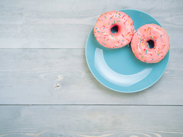 Top view of two pink donuts on gray wooden background with copy space. Colorful donuts on plate with copyspace. Glazed doughnuts with sprinkles on grey wooden table. Smile sign, good morning concept - 写真・画像