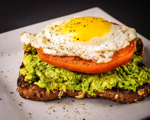 Avocado toast on white plate made with whole grain bread, sunny side up fired egg, and a sliced tomato. - Foto, afbeelding