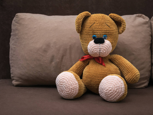 Ένα μεγάλο πλεκτό αρκουδάκι με κόκκινη κορδέλα σε μαλακό καναπέ. - Φωτογραφία, εικόνα