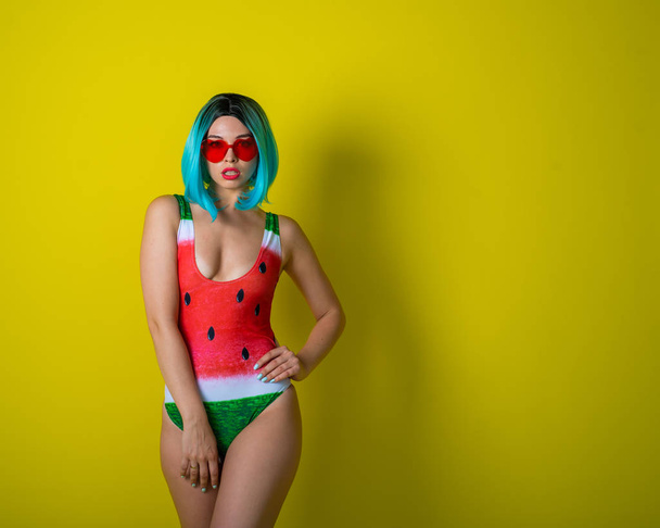 Portrait of a woman in a swimsuit with a picture of a watermelon and red sunglasses in the shape of hearts. Stylish girl in a colored short wig posing in the studio on a yellow background. - Photo, Image