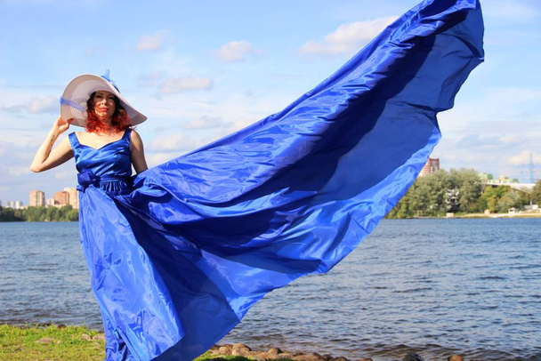 Nehir boyunca yürü. Uzun mavi elbisesi içinde kırmızı bukleli bir kadın rüzgarda savruluyor ve geniş kenarlı beyaz şapkası var. - Fotoğraf, Görsel