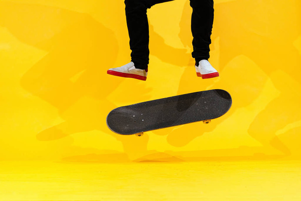 Deskorolkarz wykonujący trik deskorolkowy - kopnięcie w beton. Sportowiec olimpijski ćwiczący skok na żółtym tle w studio, przygotowujący się do zawodów. Sporty ekstremalne, kultura młodzieży - Zdjęcie, obraz