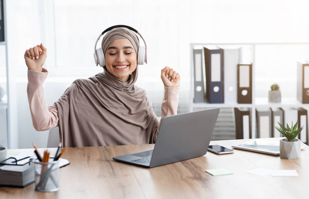 Joyeuse femme d'affaires musulmane écoutant de la musique dans des écouteurs sans fil, assise sur le lieu de travail
 - Photo, image