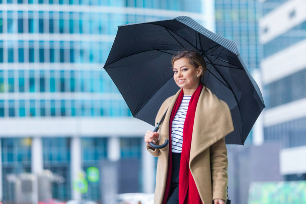 Rain. Outdoors lifestyle fashion portrait of stunning brunette girl. Walking on the city street. Going shopping. Wearing stylish white fitted coat, red neckscarf, black umbrella cane. Business woman. - Foto, afbeelding