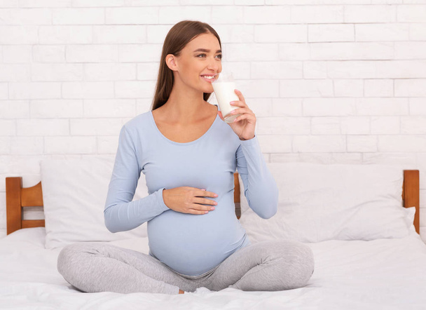 Радостная беременная женщина пьет молоко, сидя в спальне
 - Фото, изображение