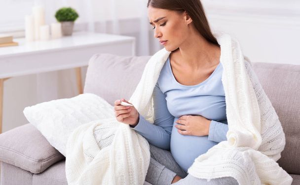 Несчастная беременная женщина измеряет температуру, сидя дома на диване
 - Фото, изображение