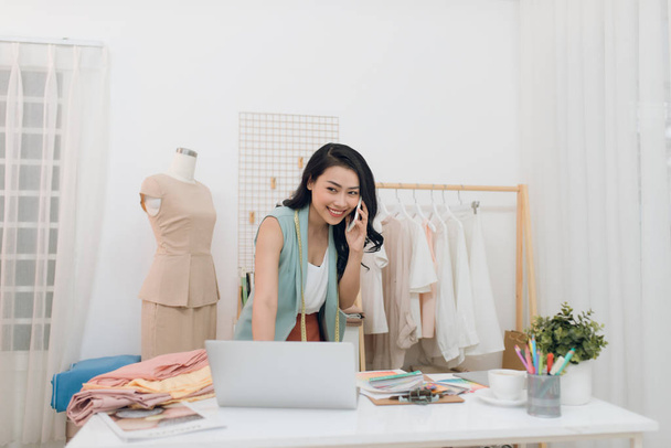 Jovem designer de moda asiática / negócios usando laptop e chamando em seu estúdio
 - Foto, Imagem