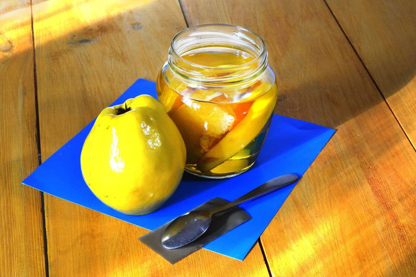 Sobre un fondo azul, sobre la mesa hay un frasco de jalea de membrillo, junto a él hay una fruta de membrillo y una cuchara pequeña yace
. - Foto, imagen