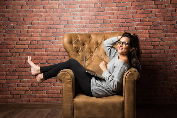 Indian girl with laptop sitting on wing chair against red brick wall - Photo, Image