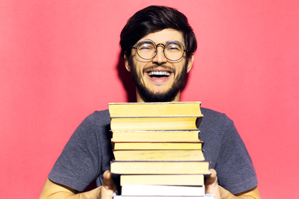 Portrait of young happy man, holding bunch of books, wearing round eyeglasses on background of coral pink color. - Fotoğraf, Görsel