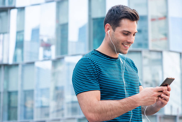 Side portrait of smiling young man listening to music with cellphone and earphones in city - Photo, image