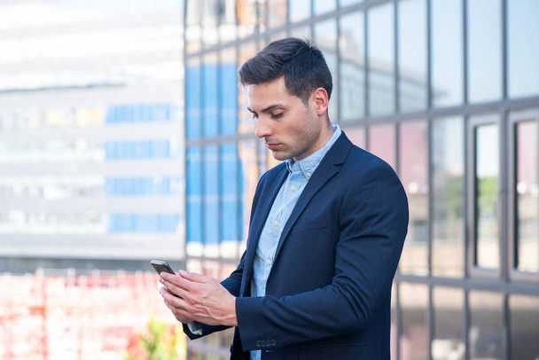 Retrato lateral de un joven empresario de pie en la ciudad mirando el teléfono móvil
 - Foto, Imagen