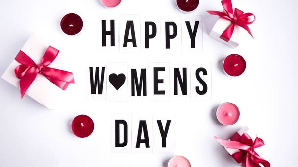 Onnellinen naisten päivä teksti valkoisella pöydällä taustalla, lahjapakkauksilla ja kynttilöillä
 - Valokuva, kuva