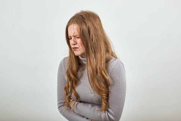 young red haired woman in gray sweater tilted her body from the pain in stomach - Photo, Image