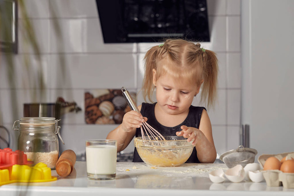 A kislány édes tortát készít liszttel, tejjel, székeken ülve egy modern konyha asztalánál. A lány habverőt tart, tojást keverget egy tálban, palacsintát készít. Kis szakács. - Fotó, kép