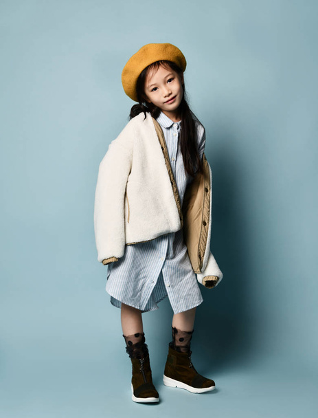 Little asian schoolgirl in shirt dress, double sided jacket, brown beret, boots. Smiling, posing on blue background. Full length - Fotoğraf, Görsel