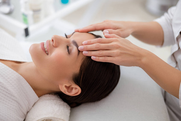 Charming young woman enjoying face massage at beauty salon - Фото, изображение