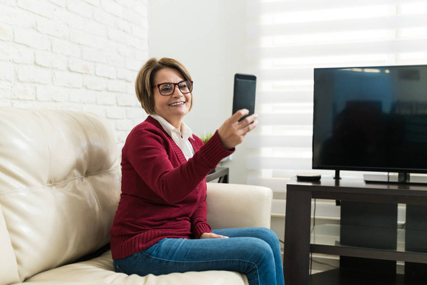 Usmívající se starší žena brát selfie přes mobilní telefon, zatímco sedí na pohovce doma - Fotografie, Obrázek