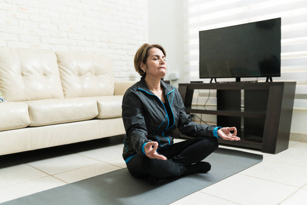 Повна довжина літньої жінки, що медитує на тренувальному килимку вдома
 - Фото, зображення