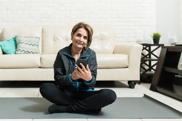 Smiling senior woman using mobile phone while sitting on exercise mat at home - Foto, afbeelding