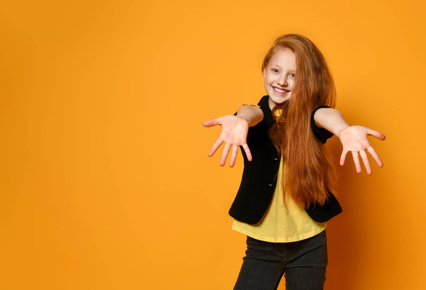 Ginger teenager in black jacket and pants, yellow t-shirt. She smiling, reaching for you, posing on orange background. Close up - 写真・画像