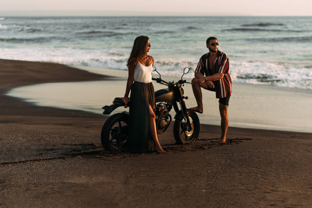 A beautiful couple on a motorbike meets the sunset by the sea. A couple in love on the beach meets the sunset. The couple is traveling on a motorcycle in Bali. Couple on a motorcycle. Lovers at sunset - Foto, afbeelding
