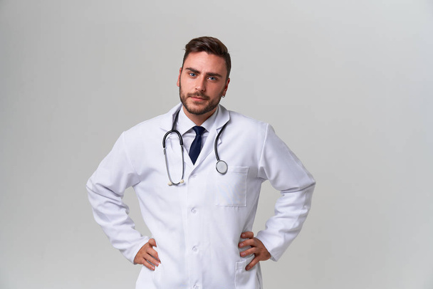 Young handsome modern doctor in a white medical gown stands in the studio on a white background. Student trainee of a medical university. - Фото, изображение