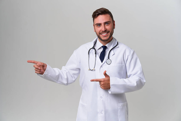 Young handsome modern doctor in a white medical gown stands in the studio on a white background. Student trainee of a medical university. - Foto, Imagen