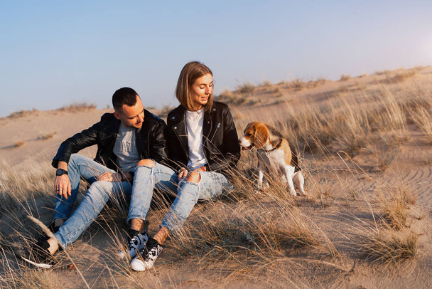 Young beautiful caucasian couple wearing leather jacket and jeans walks desert sand with Beagle dog best friend. - Fotografie, Obrázek