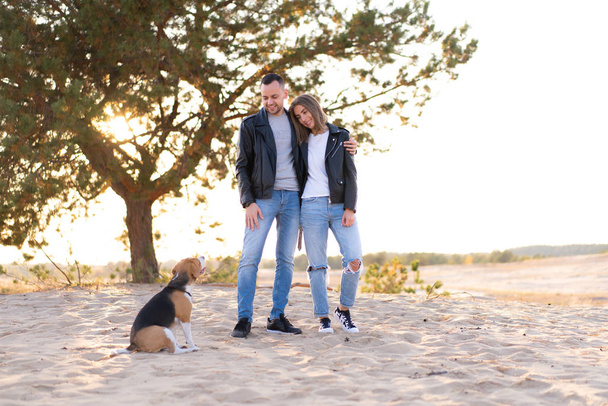 Young beautiful caucasian couple wearing leather jacket and jeans walks desert sand with Beagle dog best friend. - Foto, imagen