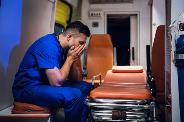 Уставший санитар в форме сидит в машине скорой помощи.
 - Фото, изображение