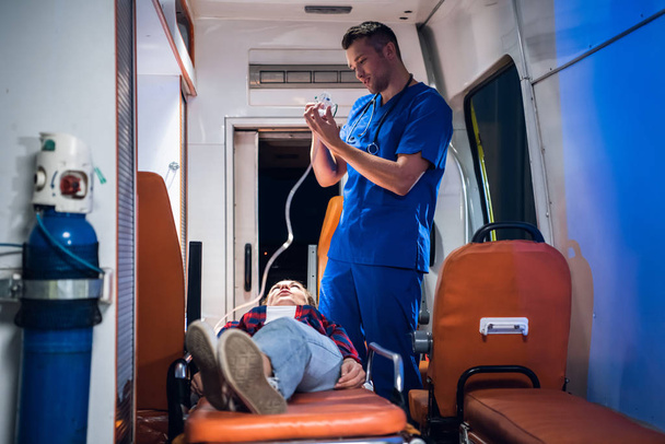 Dokter in uniform zet zuurstofmasker op vrouw liggend op brancard in de ambulance auto - Foto, afbeelding