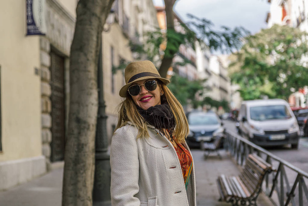 Mulher colorida atraente com casaco, chapéu, cachecol e óculos pretos sorri feliz em uma rua
 - Foto, Imagem