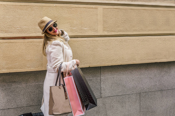 Smiling stylish blonde woman dressed in white coat walks very happy with her shopping bags - Φωτογραφία, εικόνα