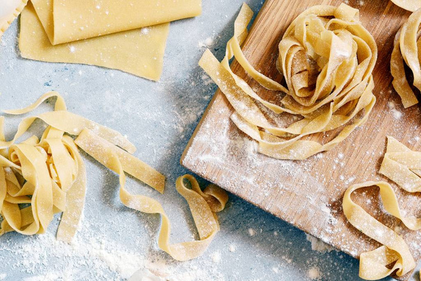 Homemade italian pasta, ravioli, fettuccine, tagliatelle on a wooden board and on a blue background. The cooking process, raw pasta. Tasty raw ravioli with ricotta and spinach,with flour on background - Photo, Image