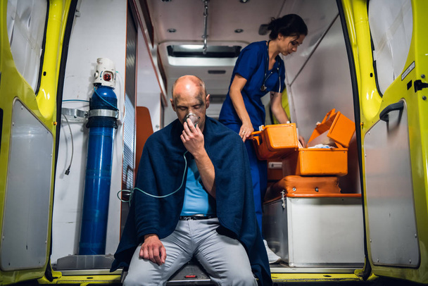 Homme respirer par masque à oxygène, jeune infirmière en uniforme ouvre sac médical dans la voiture d'ambulance
 - Photo, image