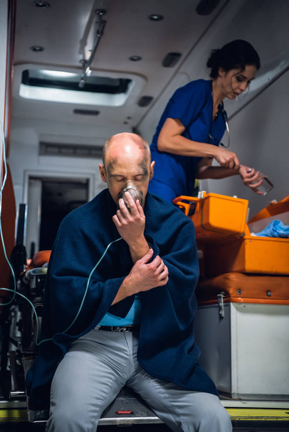 Muž v dece dýchat přes kyslíkovou masku, sestra hledat něco v lékařské tašce - Fotografie, Obrázek