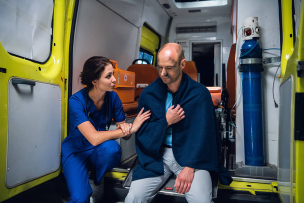 Verpleegster zit en praat vriendelijk met man in deken in ambulance auto - Foto, afbeelding